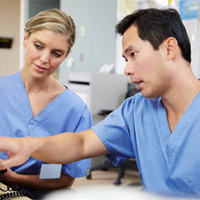 Asian man in scrubs showing White woman in scrubs something on computer at clinic nurses station.