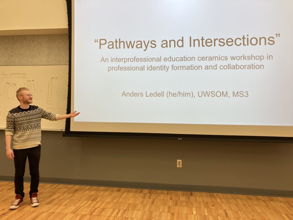 A person stands in front of a classroom with an arm directed towards a presentation that reads 